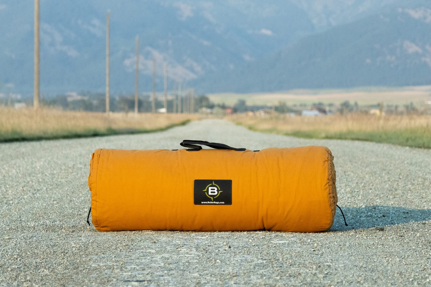 Orange roll-style duffel with a carry handle, sitting on a gravel road below mountains.