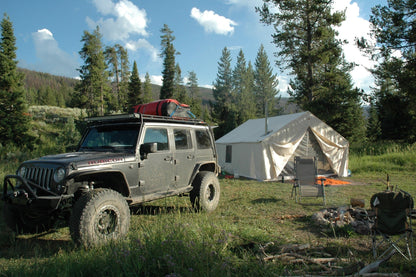 Canvas wall tent pitched in a mountain meadow beside a Jeep, with camp chairs and a fire ring.