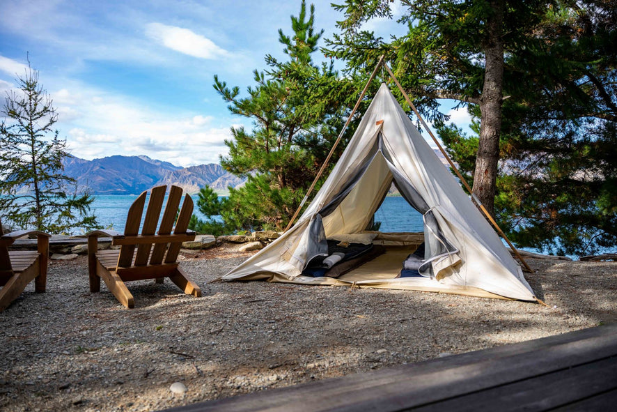 Canvas cowboy tipi open toward a lake and mountains, wooden chairs on the gravel beside it.