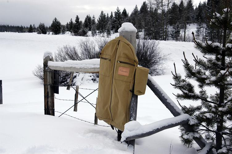 Tan canvas garment bag with zippered pockets hanging on a snowy fence rail in a pine clearing.