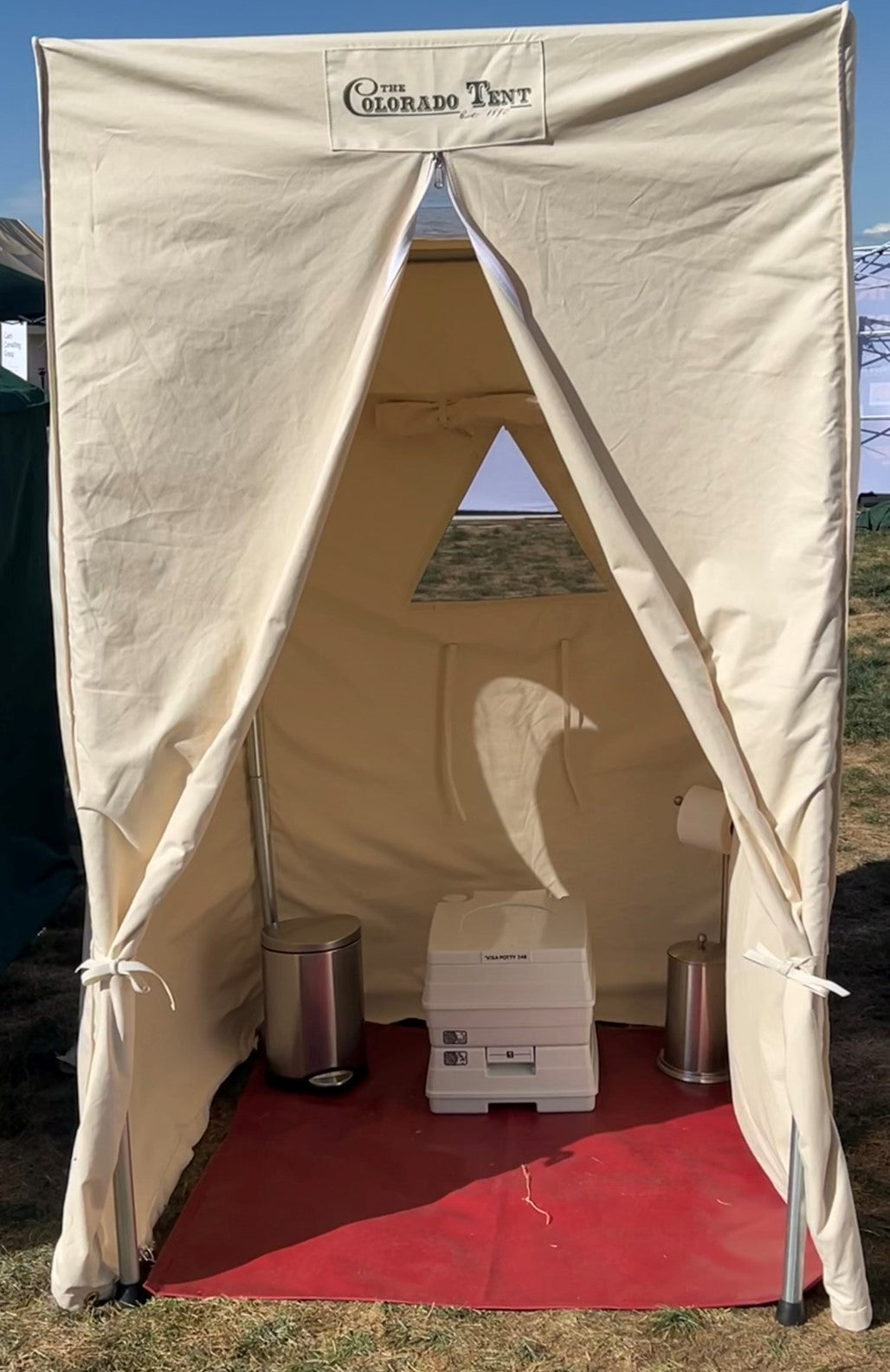 Canvas privacy tent with tied-back door flaps, a portable toilet and two trash cans inside.