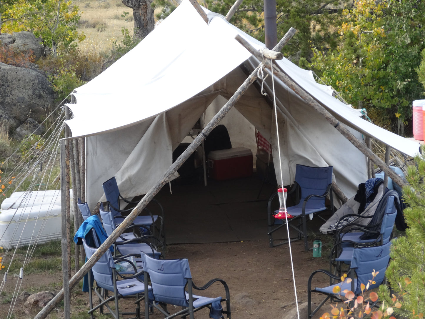 Canvas wall tent under a fly on a pole frame, camp chairs and a cooler set up at the open door.