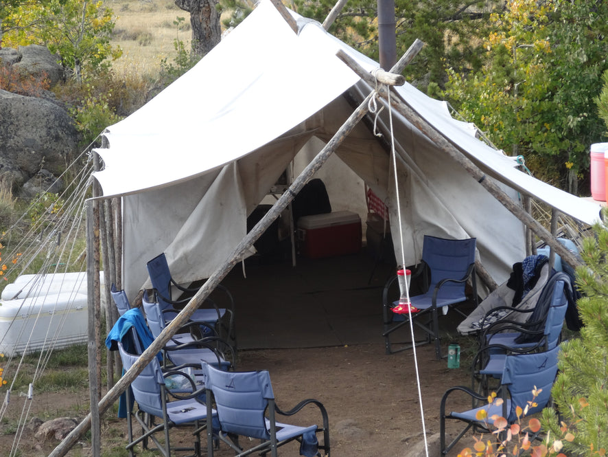 Canvas wall tent under a fly on a pole frame, camp chairs and a cooler set up at the open door.