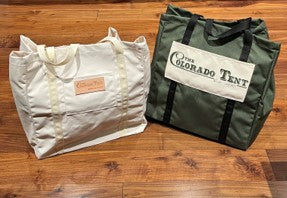 Two canvas totes on a wood floor, one natural with rope handles and one green printed Colorado Tent.