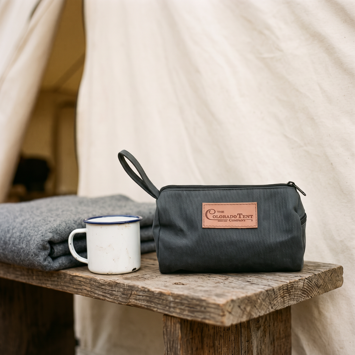 Dark canvas dopp kit with a leather Colorado Tent Company patch, on a bench by a blanket and mug.