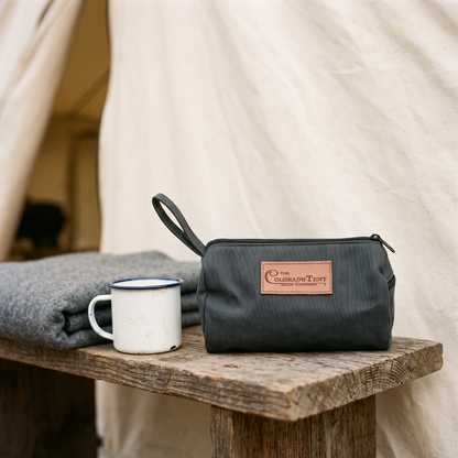 Dark canvas dopp kit with a leather Colorado Tent Company patch, on a bench by a blanket and mug.