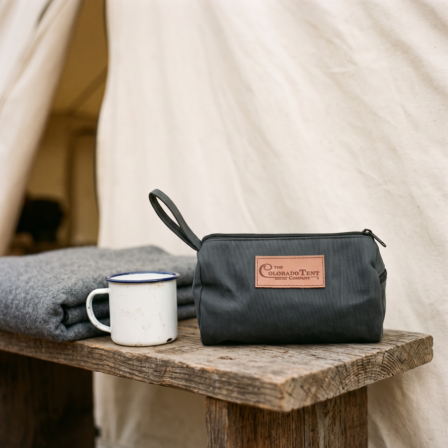 Dark canvas dopp kit with a leather Colorado Tent Company patch, on a bench by a blanket and mug.