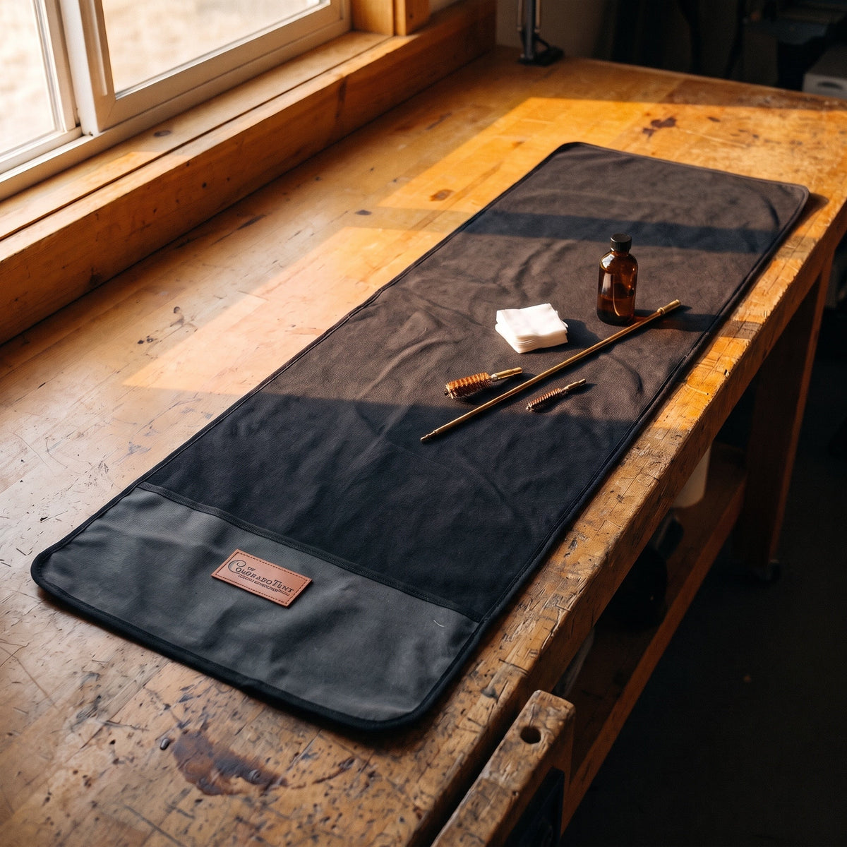 Gun Cleaning Mat in charcoal waxed canvas on a wooden workbench with a cleaning rod, bore brushes, oil, and patches