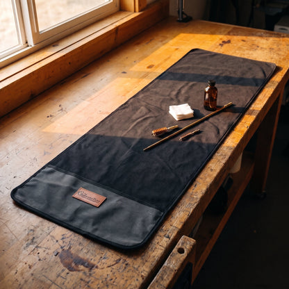 Gun Cleaning Mat in charcoal waxed canvas on a wooden workbench with a cleaning rod, bore brushes, oil, and patches