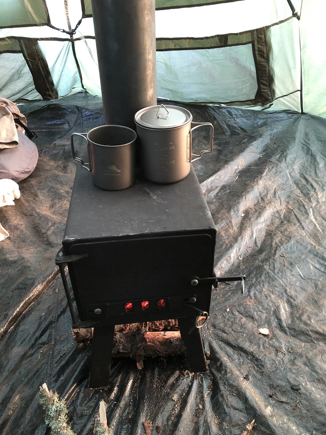 Base Camp Wood Stove by North Woods Fabrication, burning in a tent with mugs on the cooktop