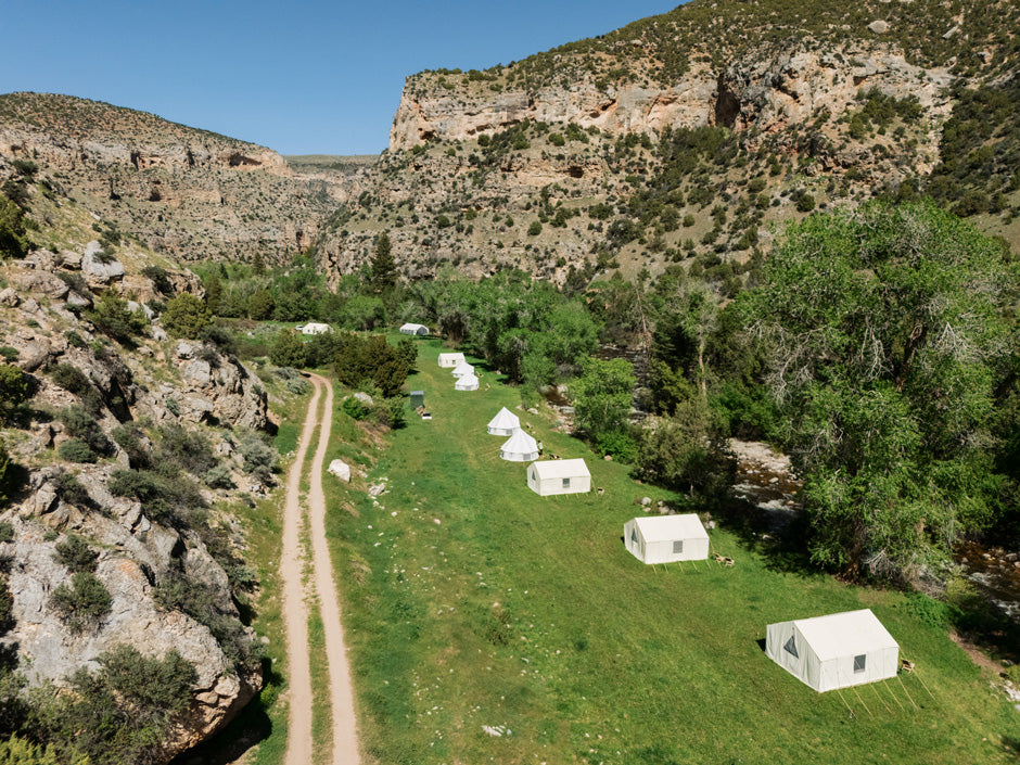 Line of canvas wall tents along a green canyon floor beside a dirt two-track road.
