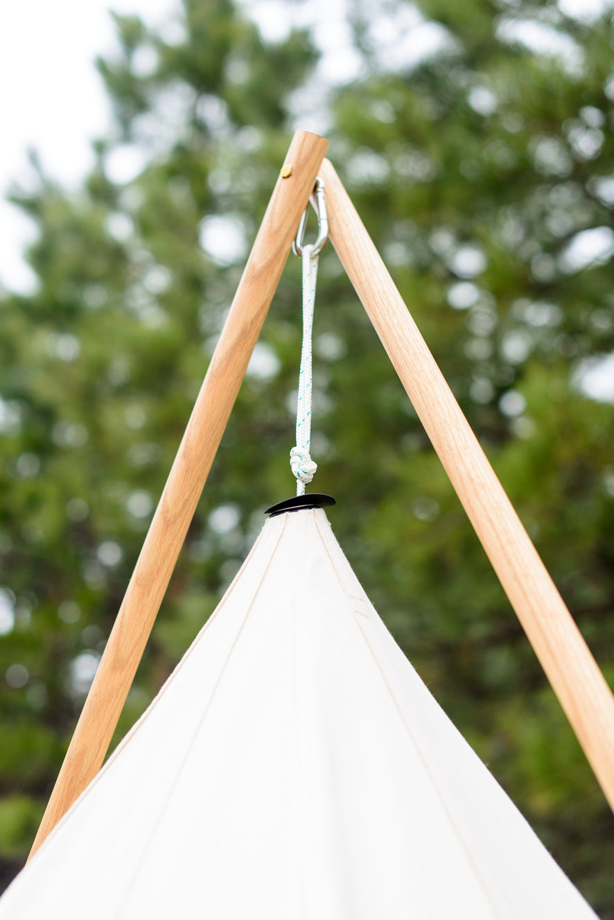 Two wooden poles crossed above a canvas tipi peak, joined by a carabiner and rope, pines behind.
